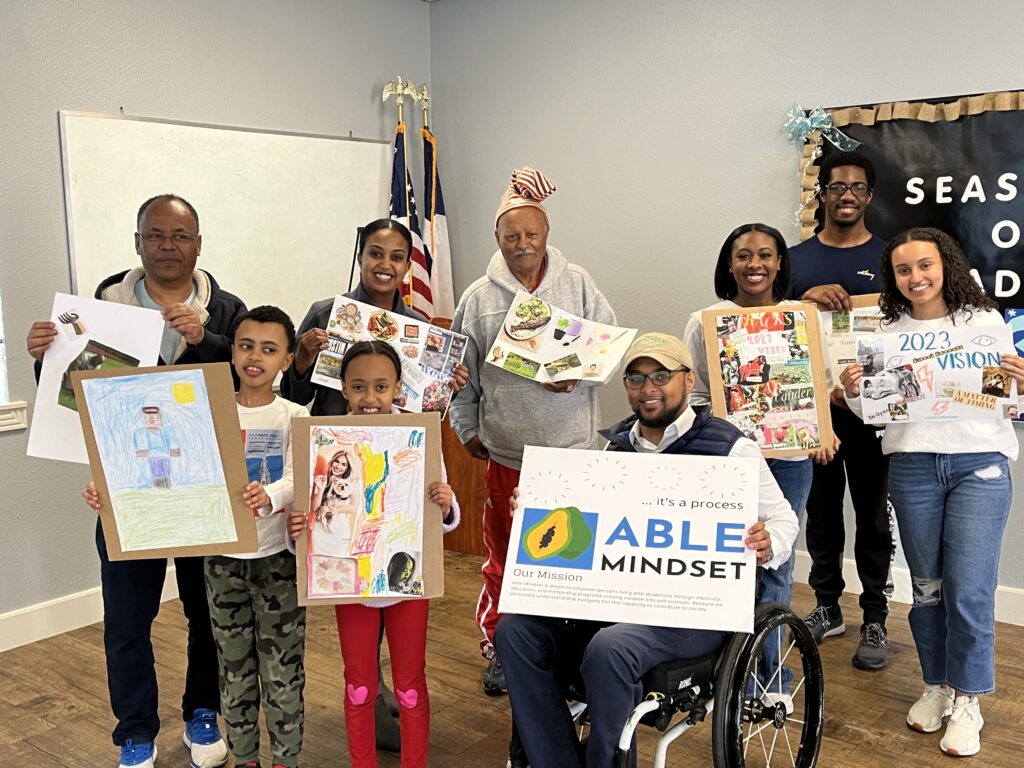 Addis Gonte of Able Mindset with a group of people holding pieces of artwork at an Able Mindset community event in Dallas Texas. Volunteer to support Able Mindset's work today